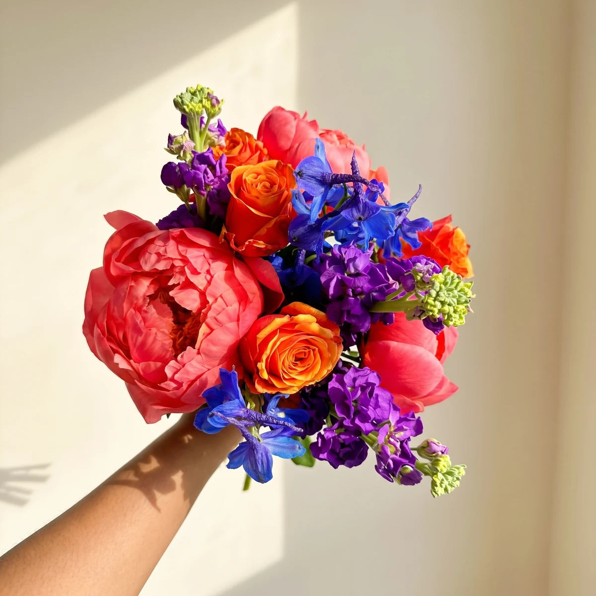 Hand holding up a bright bouquet of coral peonies, orange roses, blue delphinium and purple stocks in sunlight