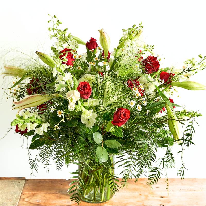 Flower arrangement of red roses, greenery, and white flowers in a clear vase on a wooden surface.