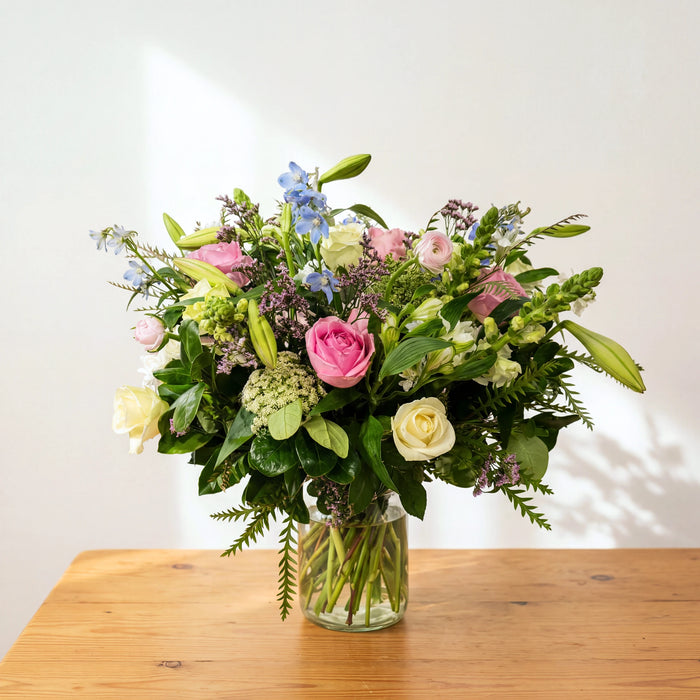 English Garden pastel flowers in a glass vase with pink roses, blue delphiniums and lily buds on a wooden table