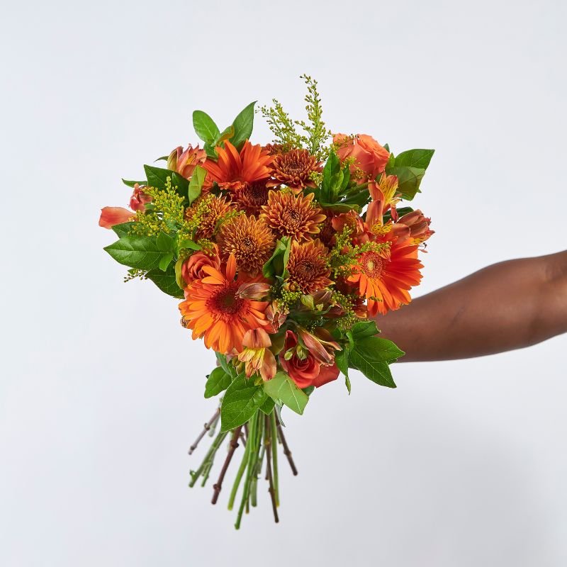 Hand holding the Firecracker Flower Bouquet featuring vibrant orange gerberas, chrysanthemums, alstroemeria, and lush greenery.
