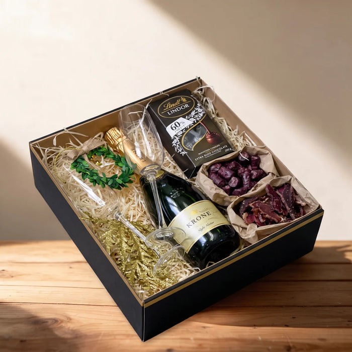Open black gift box with sparkling wine, two champagne flutes, dark chocolate, biltong and droe wors on a pine table, Fabulous Flowers South Africa