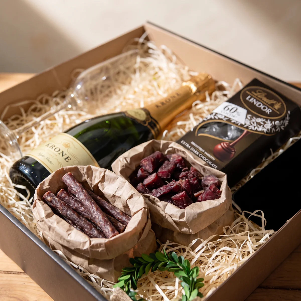 Droe wors and sliced biltong in brown paper bags beside a bottle of sparkling wine and a box of dark chocolate, Fabulous Flowers South Africa