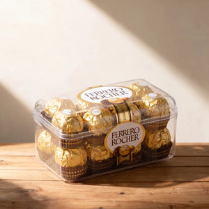 Clear box of sixteen gold-foil-wrapped hazelnut chocolates on a pine table in window light, Fabulous Flowers South Africa