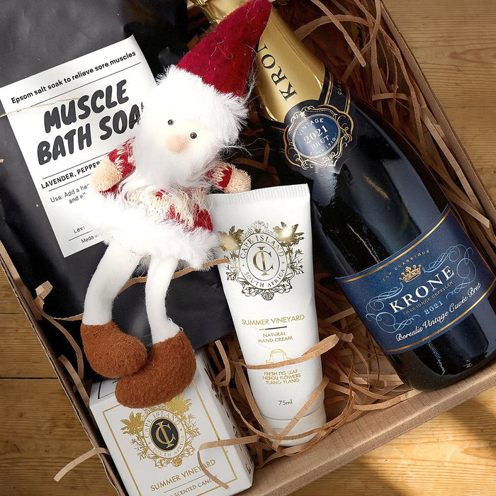Open kraft gift box from above with a bottle of Krone Borealis Vintage Cuvée Brut, a Muscle Bath Soak, a Cape Island Summer Vineyard hand cream and candle and a knitted Santa on a sunlit pine table, Fabulous Flowers South Africa