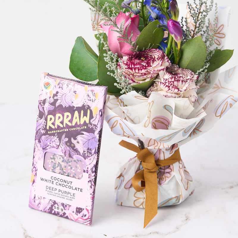 Floral Fantasy Posy - Close-up of a posy featuring pink and purple flowers wrapped in floral paper, alongside a Rrraw handcrafted coconut white chocolate bar in the 'Deep Purple' flavour.
