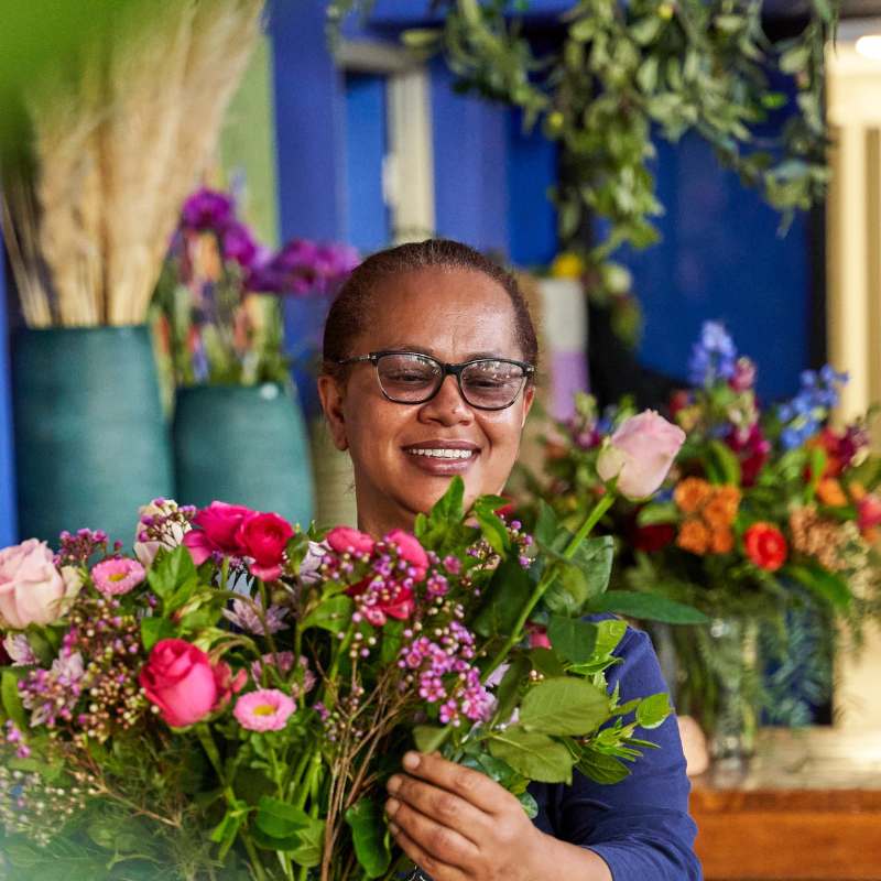 A smiling florist holding a vibrant bouquet of pink roses, daisies, and greenery, surrounded by colourful floral arrangements - Fabulous Flowers & Gifts.
