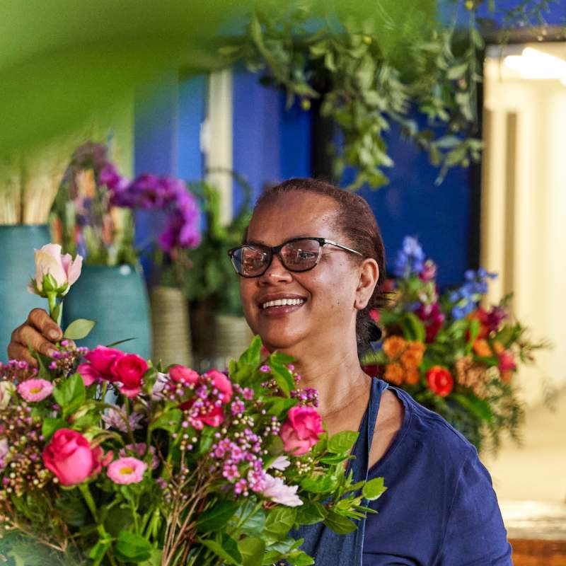 A cheerful florist arranging a bouquet of pink roses, daisies, and greenery, with colourful flowers and lush decor in the background - Fabulous Flowers & Gifts.