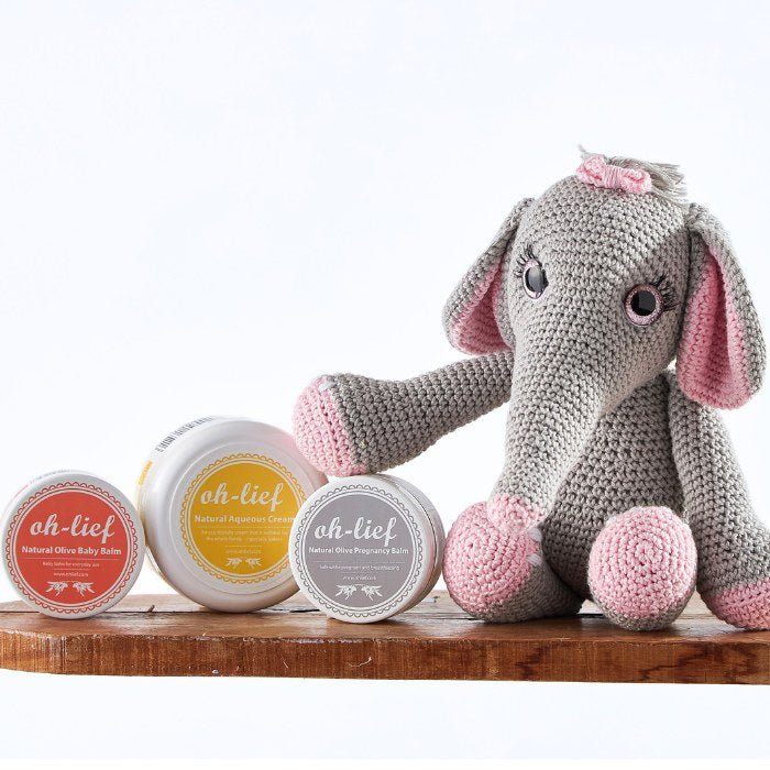 Handcrafted grey and pink crochet elephant displayed with a collection of natural baby skincare products including aqueous cream and olive balm in the Gentle Giant Skincare Bundle.