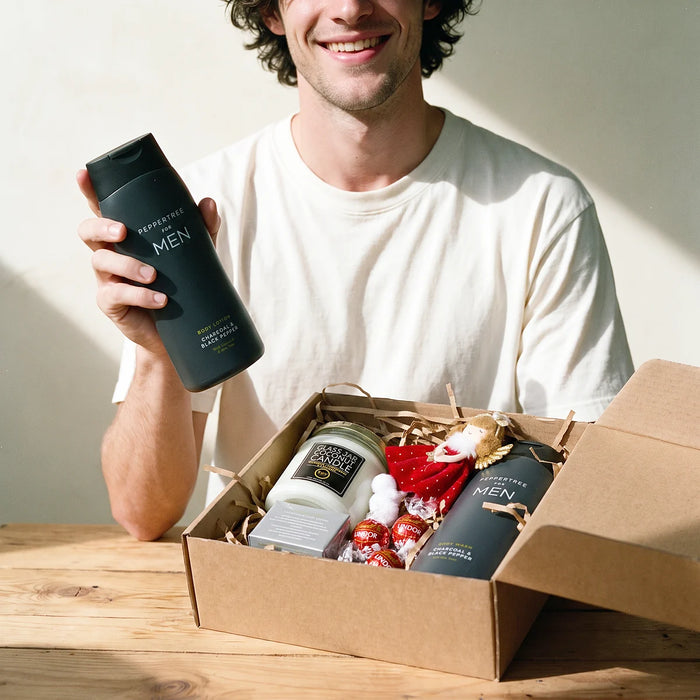 Grinning man in a white t-shirt holding up a Peppertree for Men charcoal and black pepper body lotion beside an open Christmas gift box, Fabulous Flowers South Africa