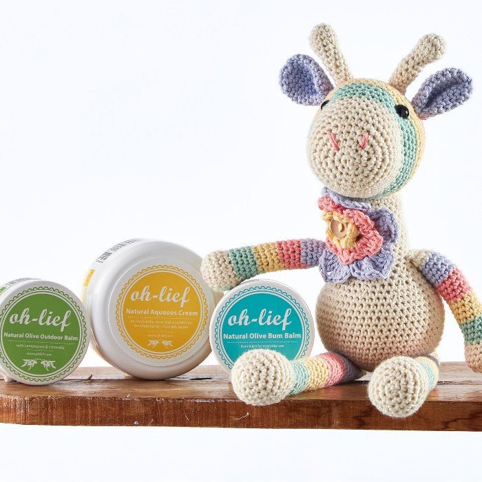 Colorful crocheted giraffe toy alongside natural baby skincare products, featuring olive bum, aqueous, and outdoor balms on a rustic wooden surface.