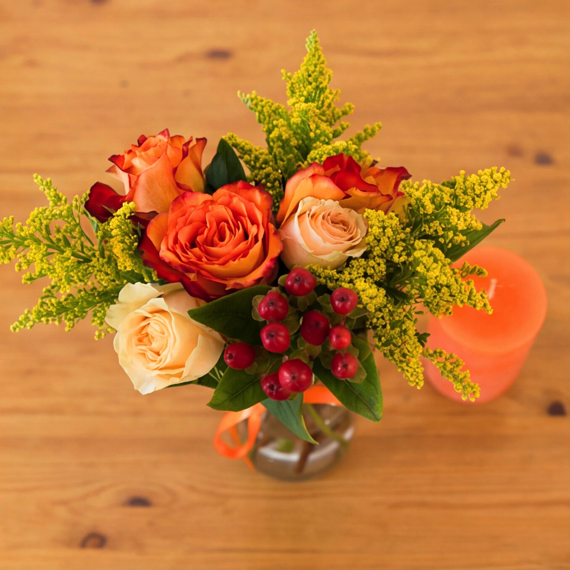Top down view of orange and cream roses arranged in a vase with berries, solidago and an orange candle