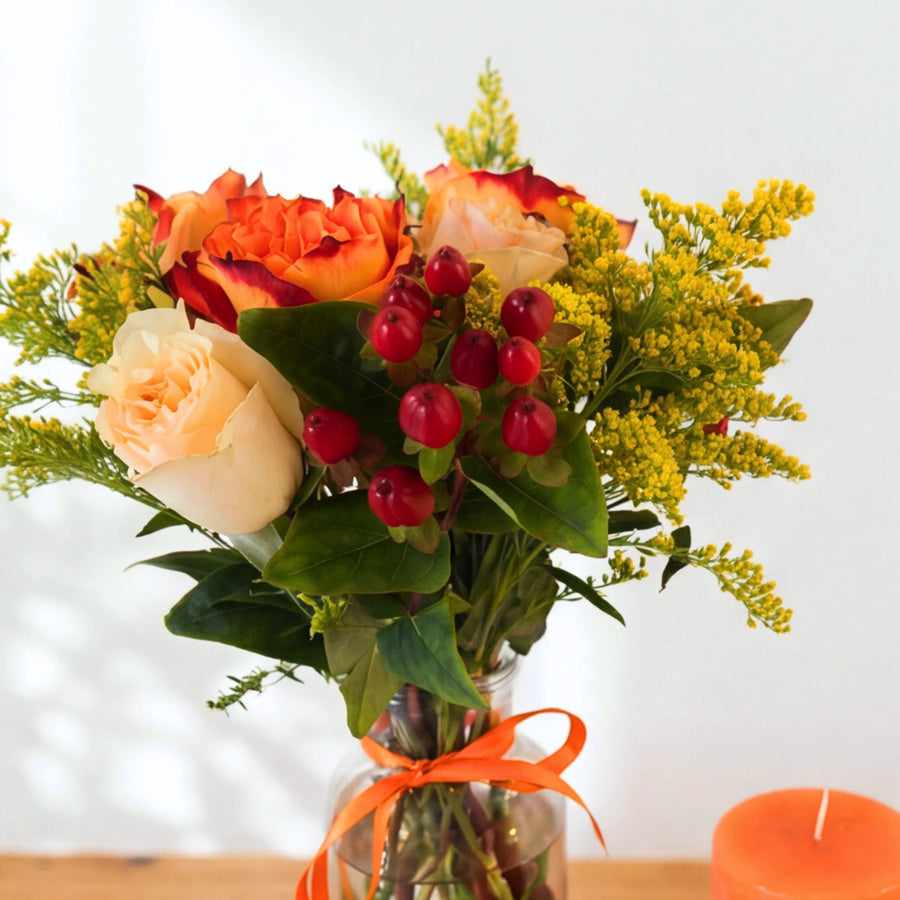 Hand-tied posy of sunset roses, cream garden roses and red berries in a glass vase with orange ribbon