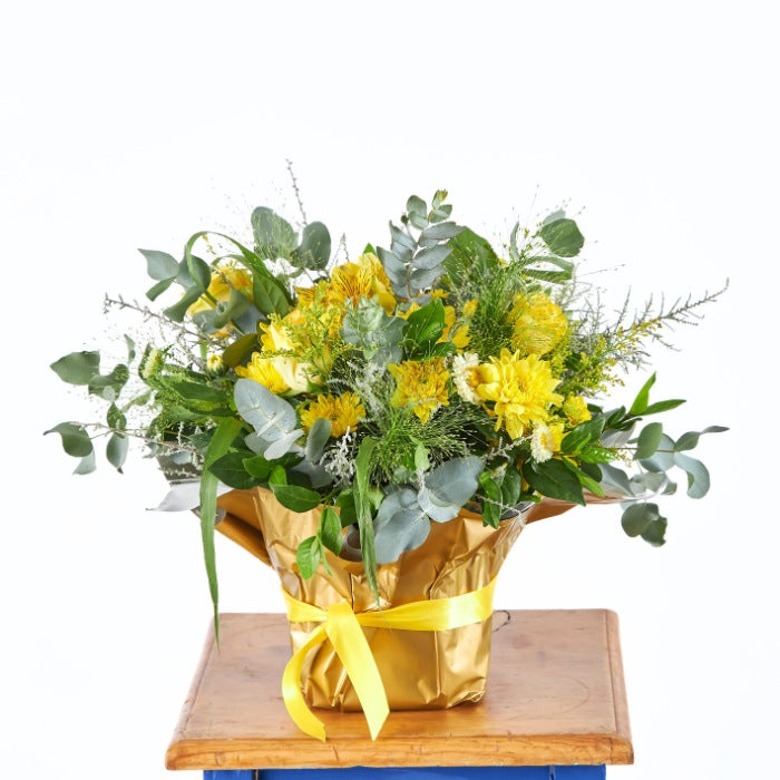 A lavish display of vibrant yellow flowers in a golden wrapped pot, including roses and wildflowers, exemplifying richness and celebration.