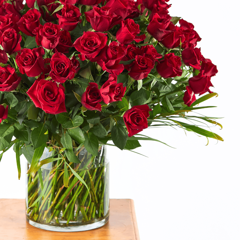 luxurious bouquet of ruby red roses in a glass vase with lush greenery, displayed on a wooden table