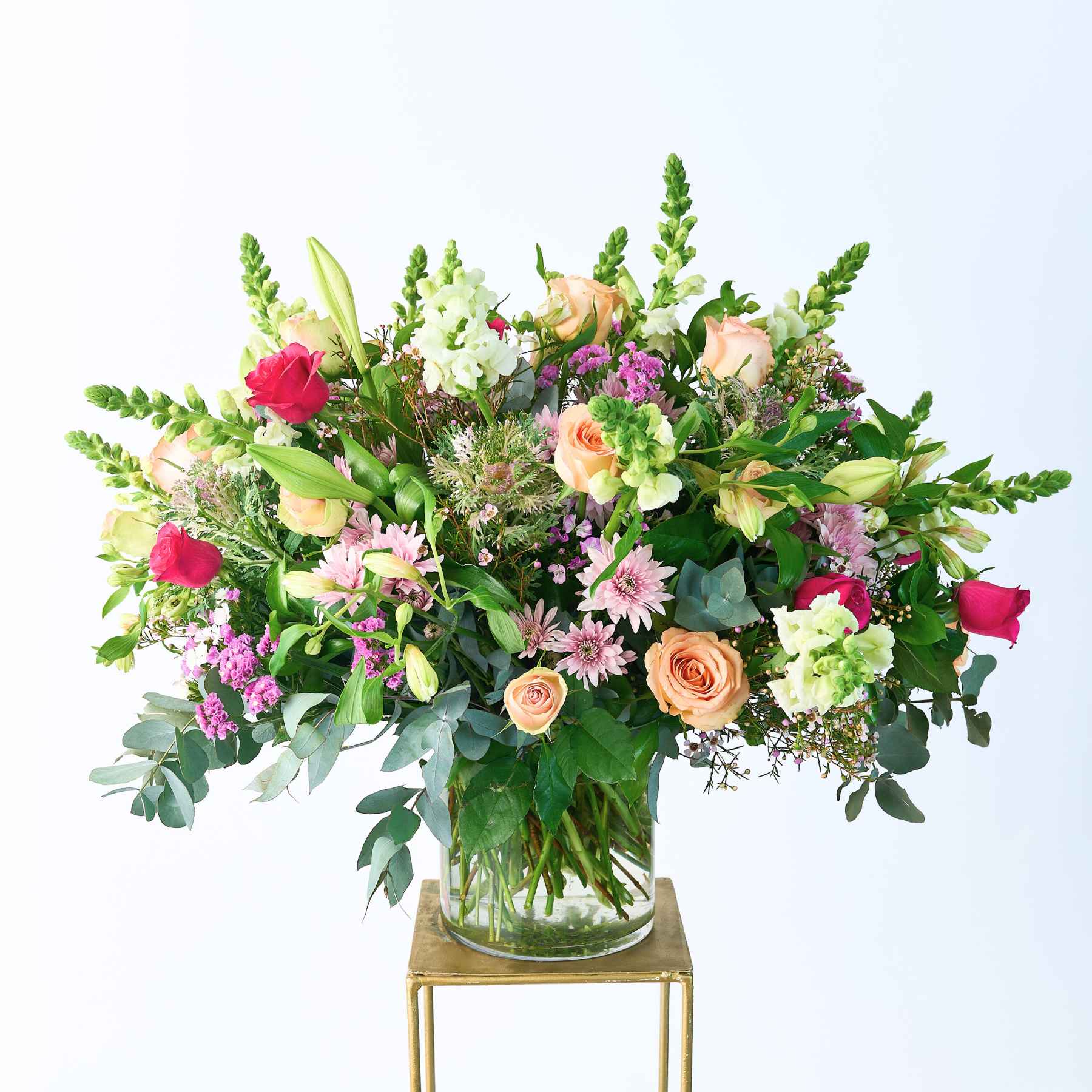 A grand and colourful flower arrangement in a glass vase, featuring a vibrant mix of roses, lilies, chrysanthemums, and greenery, creating a lively and elegant display.