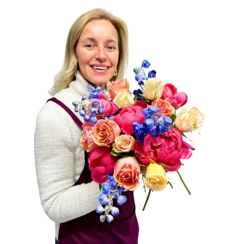 Smiling florist in white jumper and plum apron holding Heart & Bloom bouquet with coral peonies, roses and blue delphiniums.