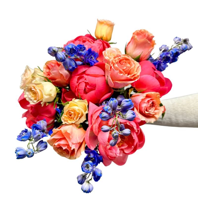 Heart & Bloom bouquet featuring vibrant coral peonies, peach and yellow roses, and blue delphiniums against white background.