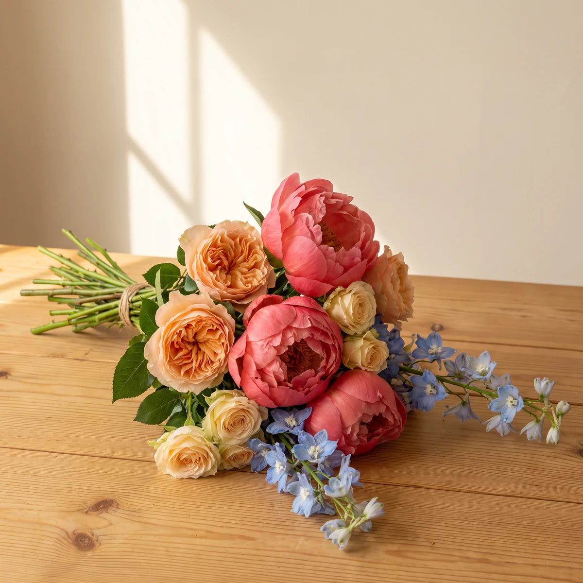 Hand-tied bouquet of coral peonies, peach and cream roses and blue delphinium lying on a wooden table in afternoon sun