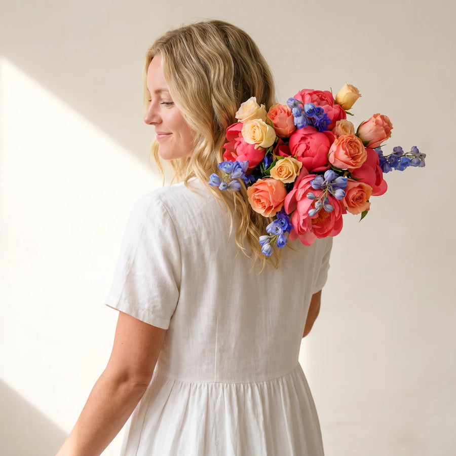 Woman with blonde hair smiling over her shoulder, carrying a bouquet of coral peonies, peach roses and blue delphinium