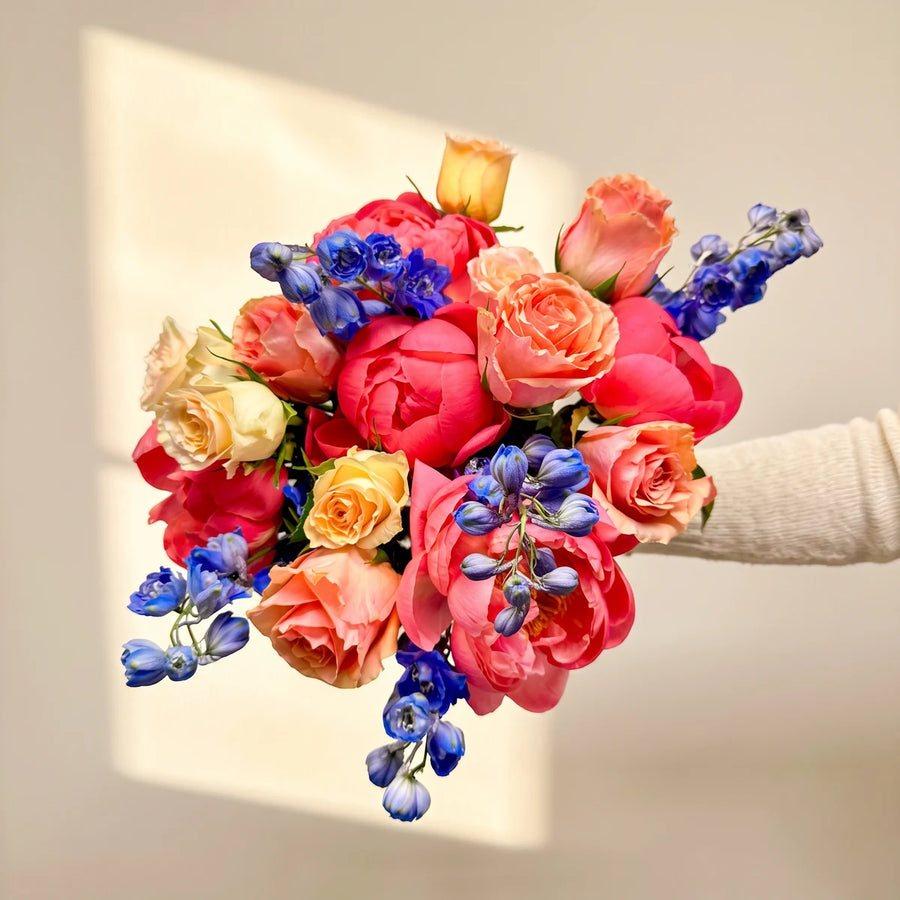 Arm holding out a bouquet of coral peonies, peach and cream roses and blue delphinium against a sunlit wall