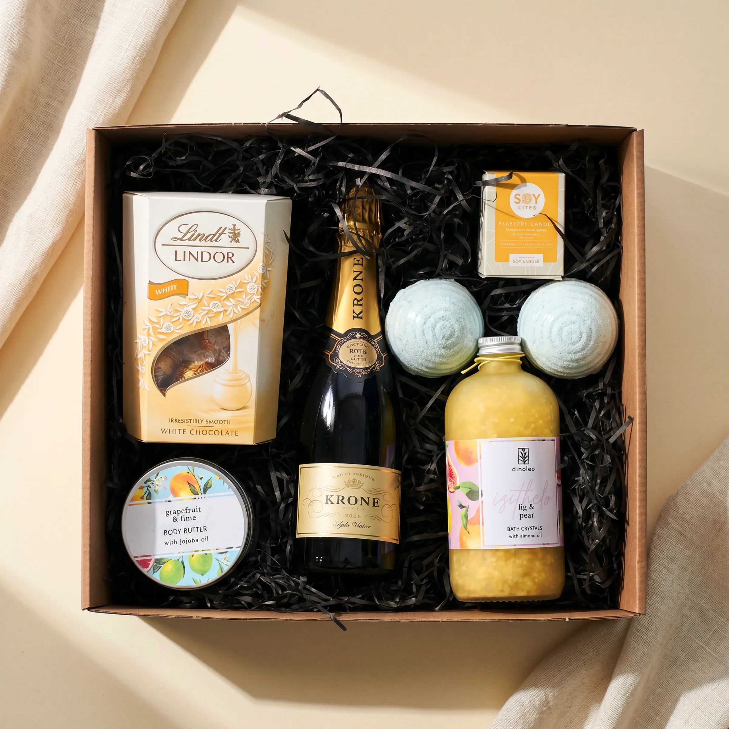 Top-down view of The Heavenly Harmony Hamper by Fabulous Flowers South Africa, Krone Cap Classique sparkling wine, Lindt Lindor White truffles, Grapefruit and Lime body butter with jojoba oil, Fig and Pear bath crystals, two bath bombs and a Soy Lites soy candle hand-packed in a kraft keepsake gift box