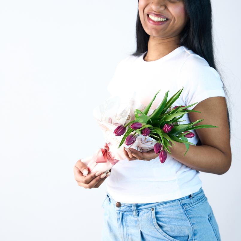 Smiling woman holding a small bouquet of purple tulips wrapped in pastel floral paper, wearing a white t-shirt and jeans.