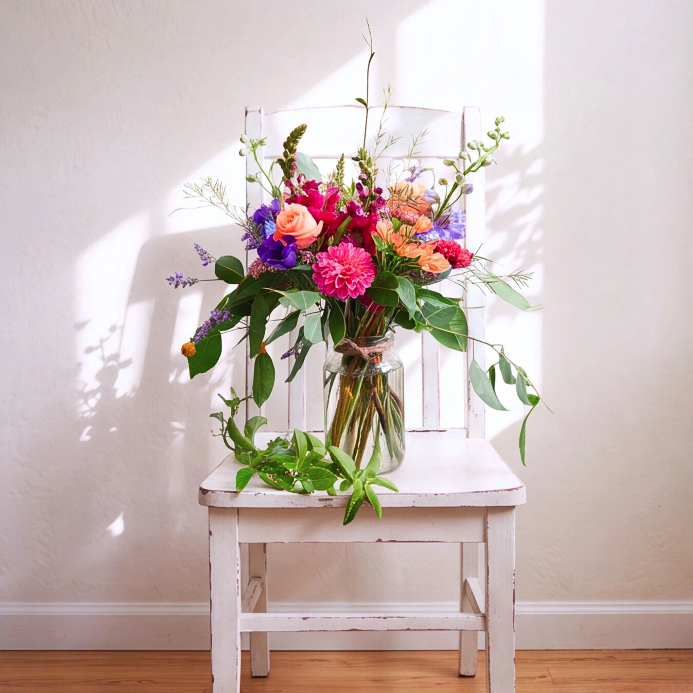 A vibrant, multicolored wildflower bouquet in a clear glass jar vase, styled on a distressed white wooden chair against a textured cream wall with soft window light shadows.