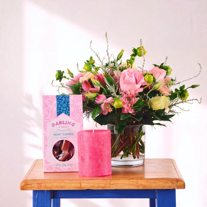 A pink candle and a pink box of Darling Mint Toffee chocolates are styled on a wooden table next to a vibrant pink flower arrangement in a glass vase against a white wall.