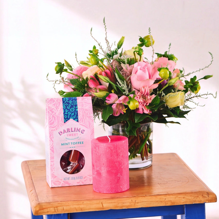 A pink box of Darling Confections Mini Toffee next to a textured hot pink pillar candle, set on a wooden stool in front of a vibrant pink and yellow flower arrangement.
