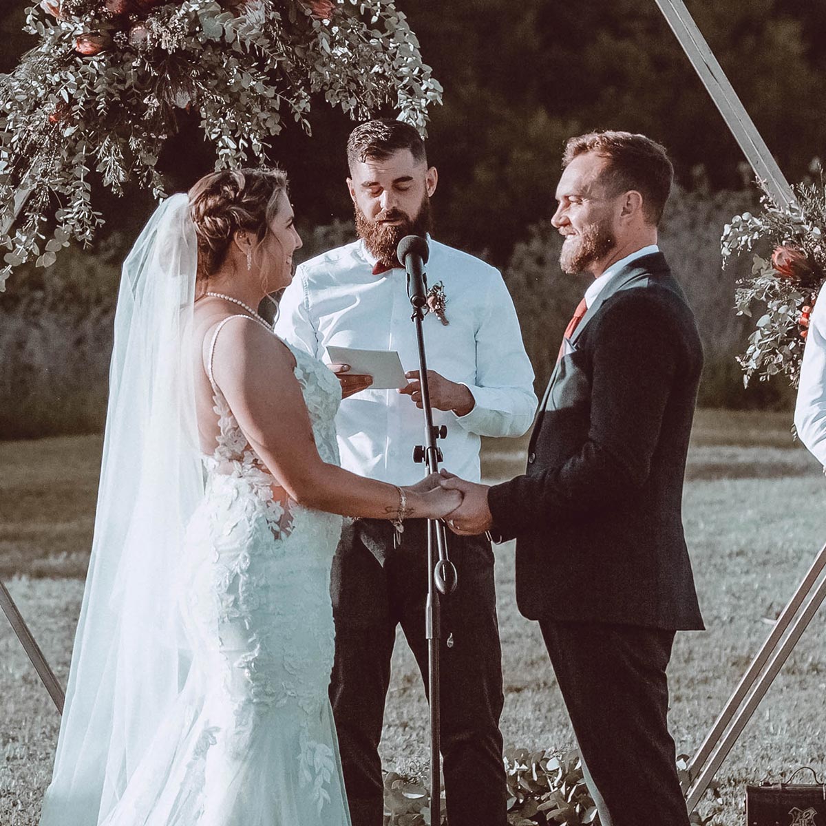 Bride Megan and groom Steve exchange vows outdoors under a floral arch, holding hands and smiling, with the officiant reading from a script. Weddings. Fabulous Flowers and Gifts.