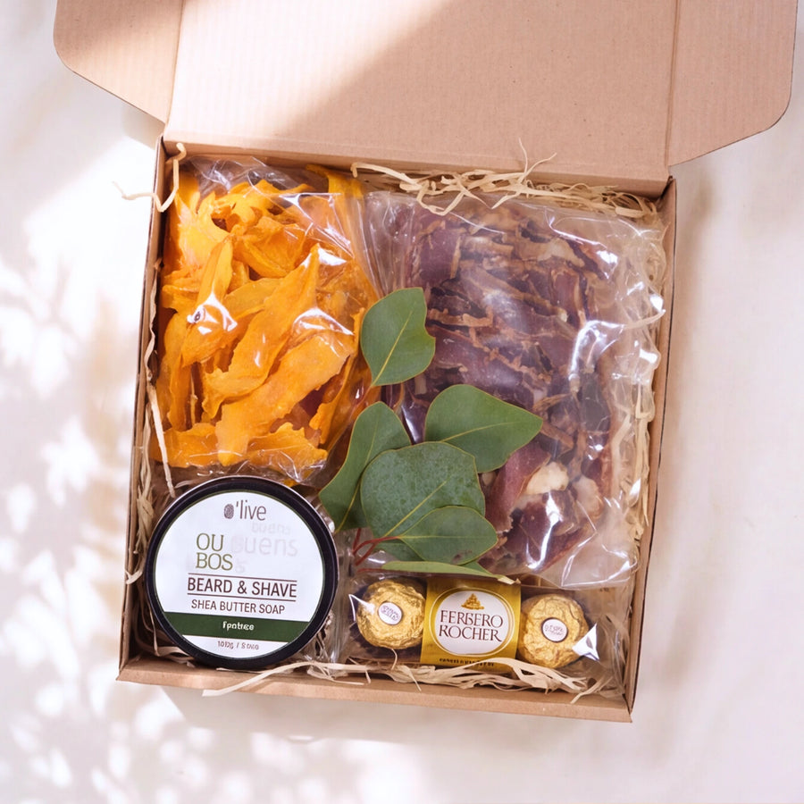 An open kraft gift box filled with beard grooming items, including dried orange and brown botanicals in clear bags, a round tin of OU Bio Beard & Shave Shea Butter Soap, and two wrapped Ferrero Rocher chocolates, arranged on shredded paper.