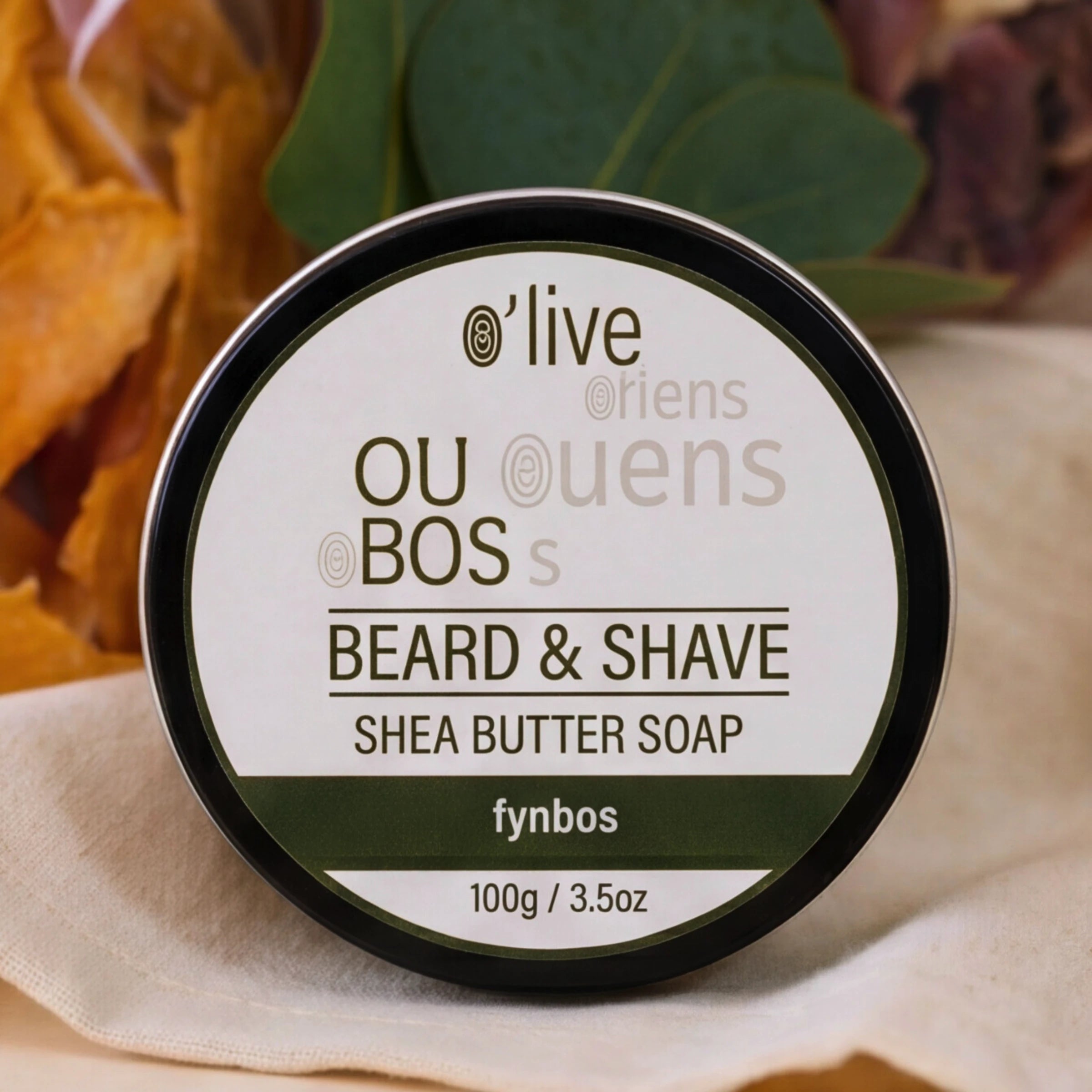 A round tin of fynbos OU BOS o'live Friends Beard & Shave Shea Butter Soap, featuring a white label with dark green and black text, set against a rustic background with dried leaves and a beige cloth.
