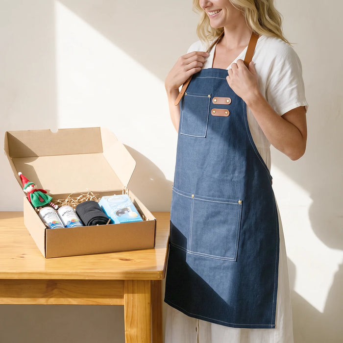 Smiling woman wearing a navy denim apron with leather straps beside an open Christmas gift box of seltzers, socks and Lindt chocolate, Fabulous Flowers South Africa