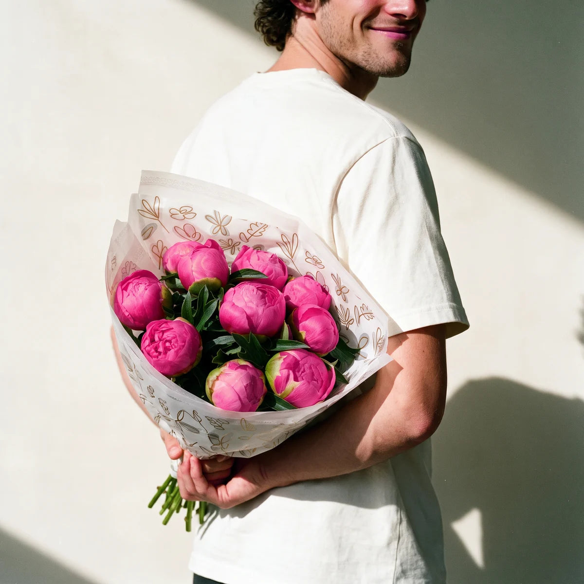 Man with curly dark hair in a white t-shirt grinning as he hides a bouquet of pink Monsieur Jules Elie peonies behind his back, Fabulous Flowers South Africa