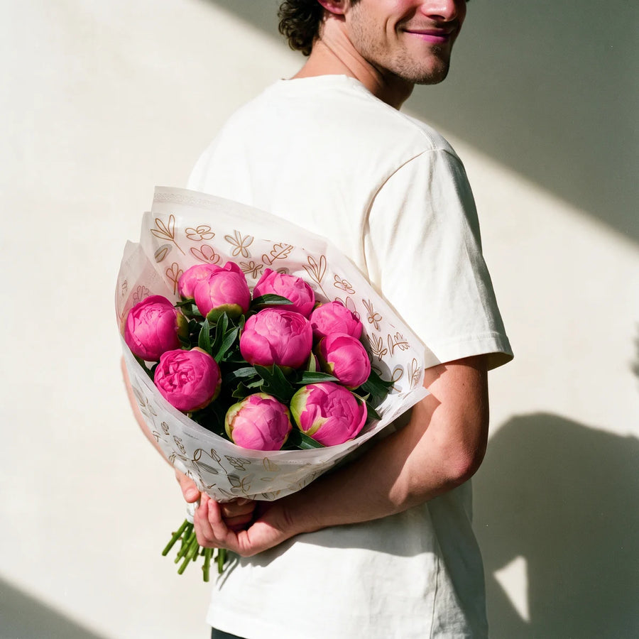 Man with curly dark hair in a white t-shirt grinning as he hides a bouquet of pink Monsieur Jules Elie peonies behind his back, Fabulous Flowers South Africa