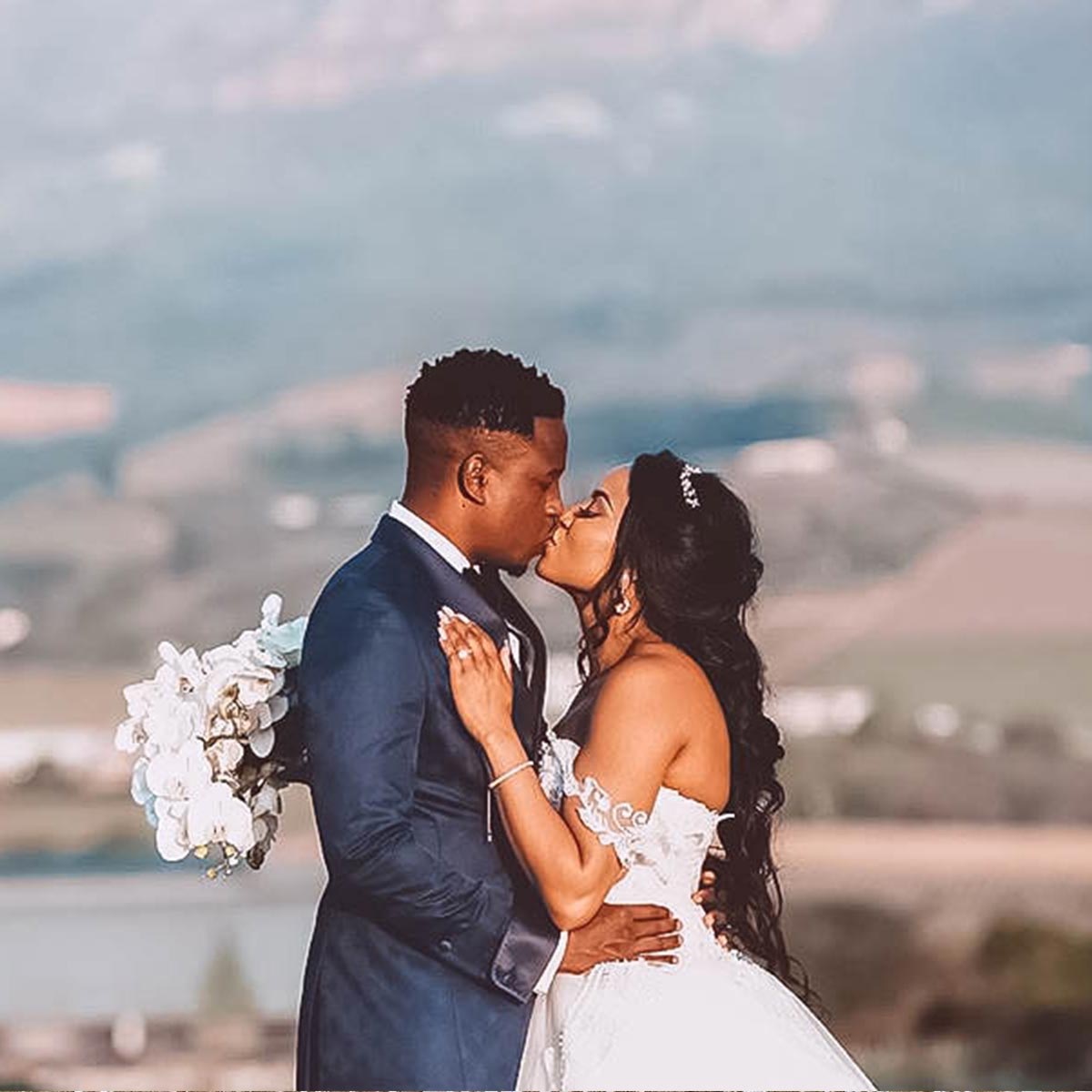 Bride Pinkie and groom Joe share a romantic kiss with a scenic valley in the background. Pinkie holds a bouquet of white flowers. Weddings. Fabulous Flowers and Gifts.