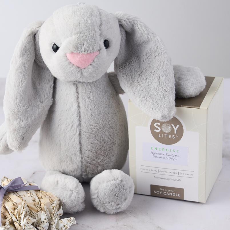 Close-up of the Pamper Bunny for Baby Boy Gift Set featuring a plush grey bunny, Soy Lites Energise candle, and Sally Williams nougat on a white surface.