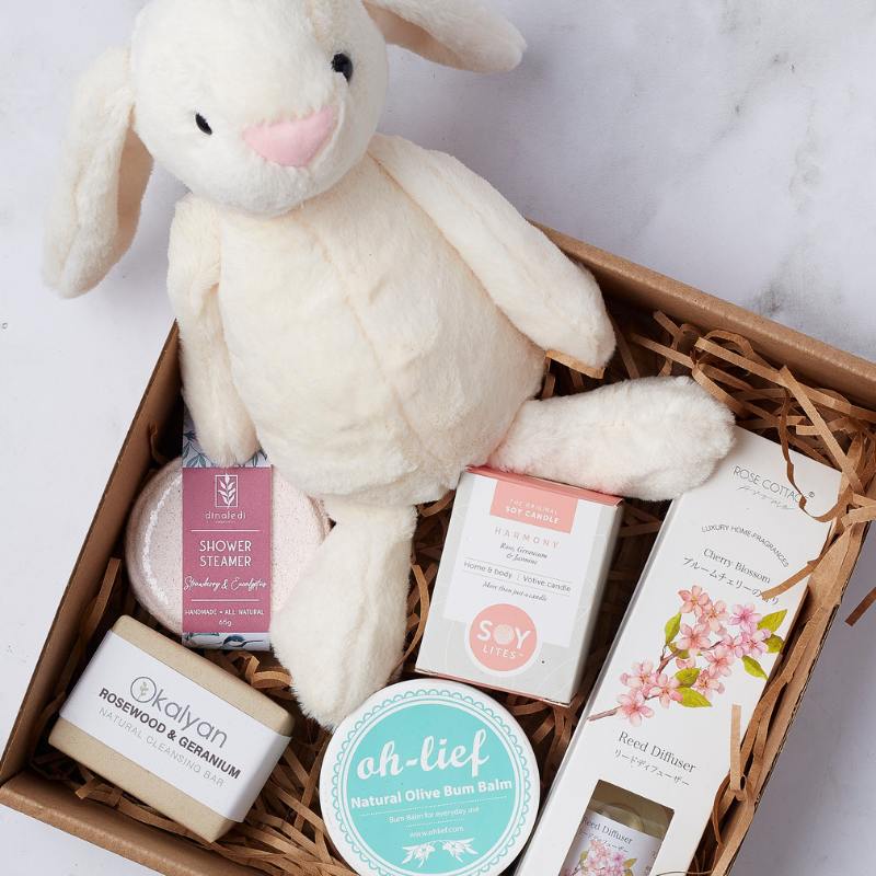 Close-up of the Pamper Bunny for Baby Girl gift box with a plush bunny toy, natural olive bum balm, shower steamer, soap bar, and reed diffuser.