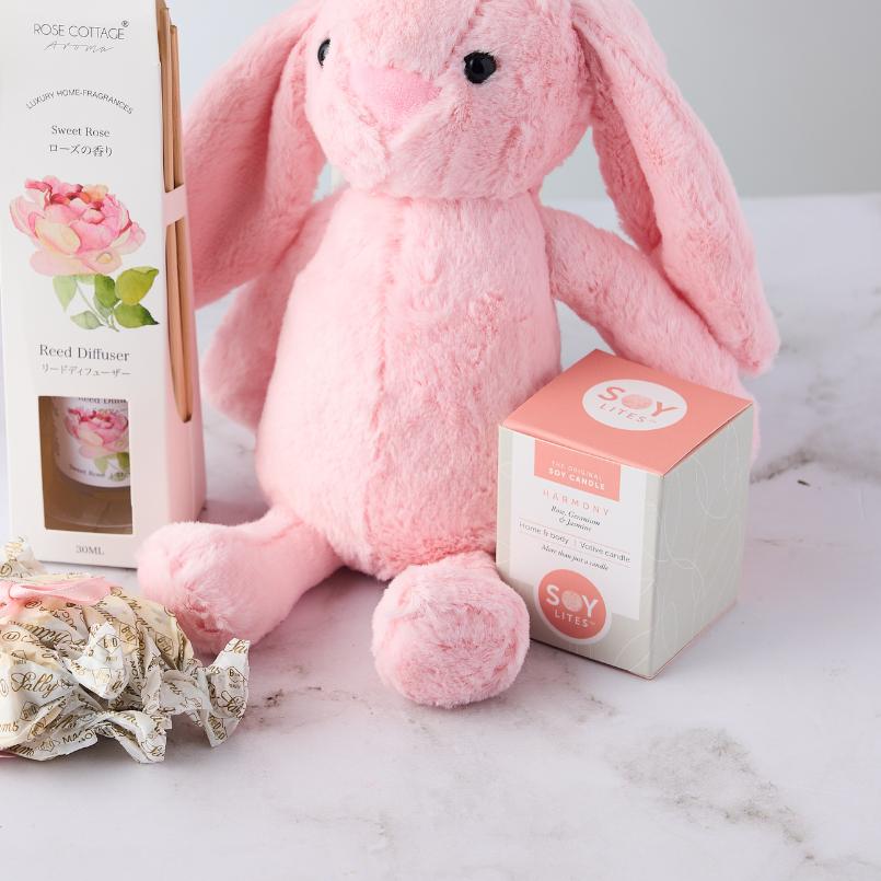 Close-up of Pamper Bunny Gift Set with a pink plush bunny, Soy Lites candle, Rose Cottage reed diffuser, and Sally Williams nougat on display.