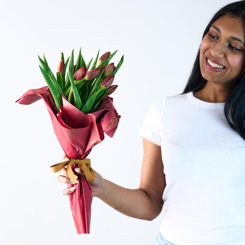 Woman smiling while holding a Passionate Embrace Tulip Bouquet with vibrant red tulips and lush green stems wrapped in red and gold paper.