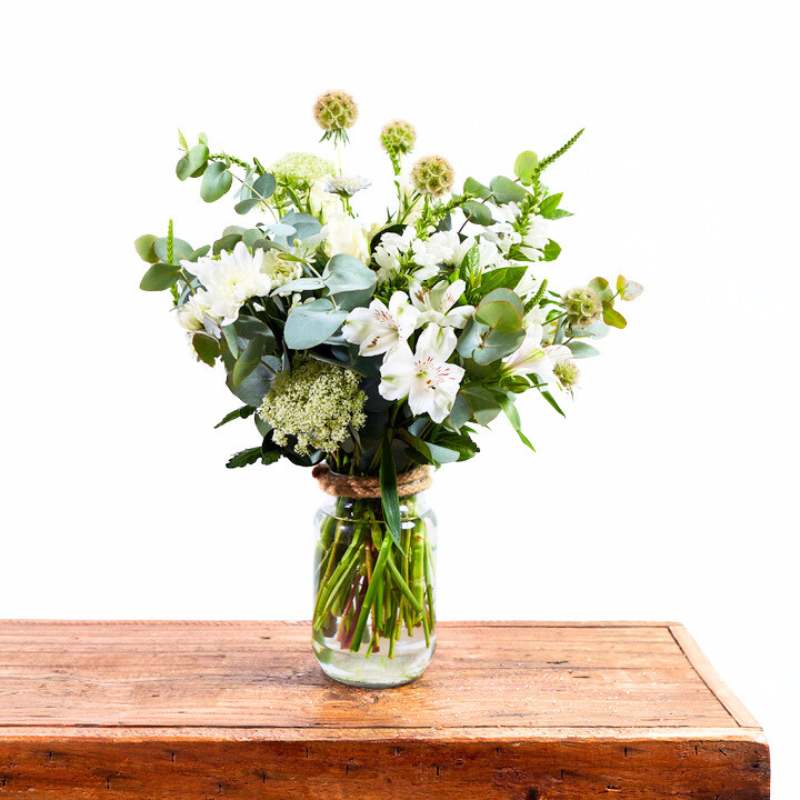 Jar arrangement of white and green flowers in a clear vase on a wooden table with a white background