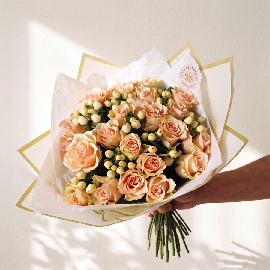 A bouquet of peach and pink roses with green stems, wrapped in white tissue paper with gold geometric borders, held by a hand against a warm off-white background with soft window shadows.