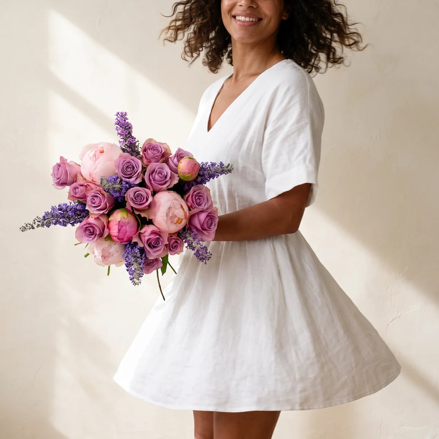 Smiling woman with curly hair in a white dress twirling with a bouquet of pink peonies and lilac roses