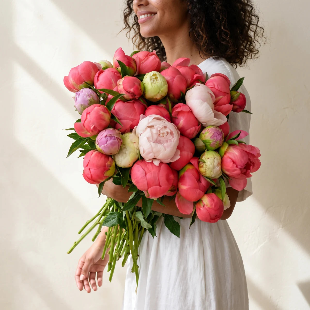Smiling woman with curly hair in a white dress carrying an armful of coral and blush peonies