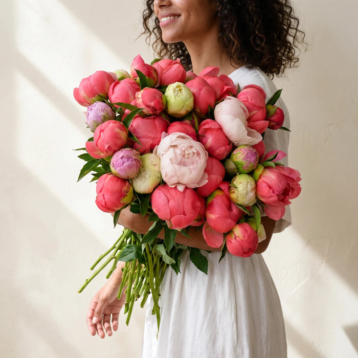 Smiling woman with curly hair in a white dress carrying an armful of coral and blush peonies