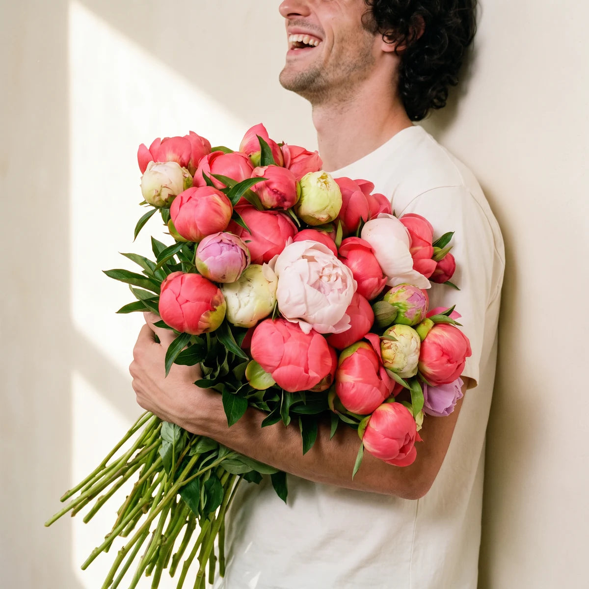 Laughing man in a white T-shirt hugging an armful of coral and pink peonies against a sunlit wall