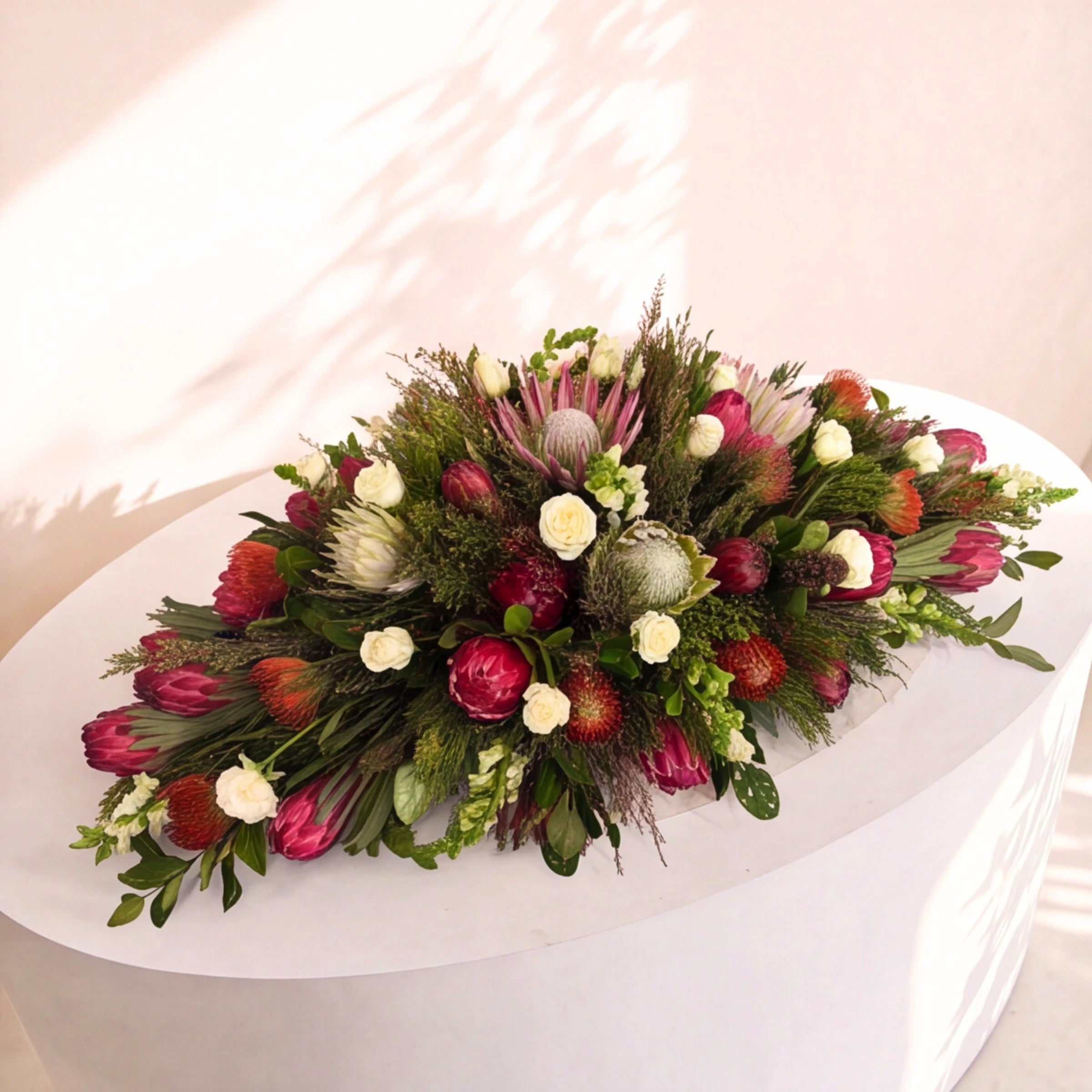 A large, lush floral arrangement featuring red protea-like flowers, green foliage, white lilies, and pinkish-purple protea blooms, displayed on a white pedestal against a softly lit beige wall with leaf shadows.