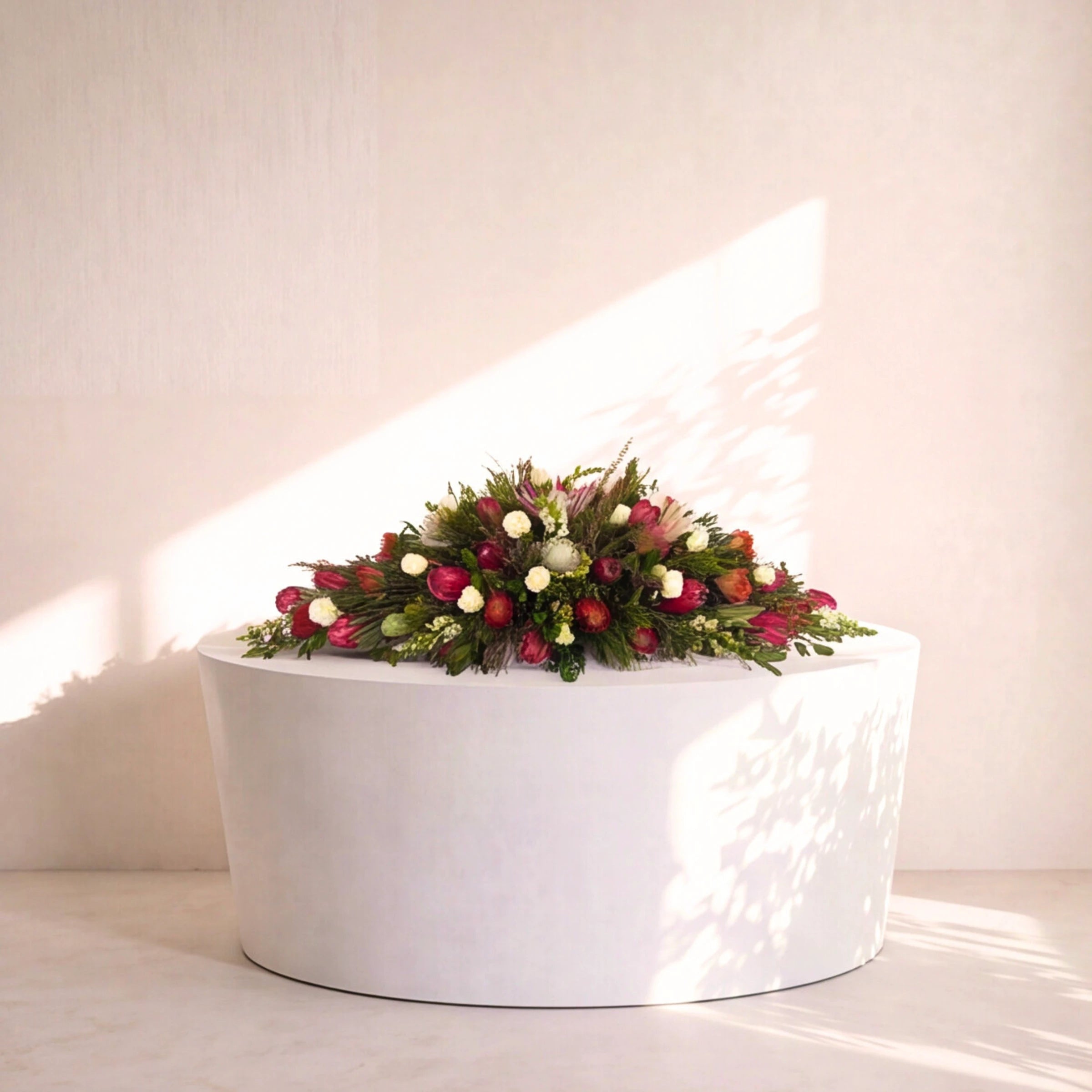 A floral arrangement with red berries, green foliage, and small white flowers displayed in a round white planter against a light beige wall with soft sunlight and leaf shadows.