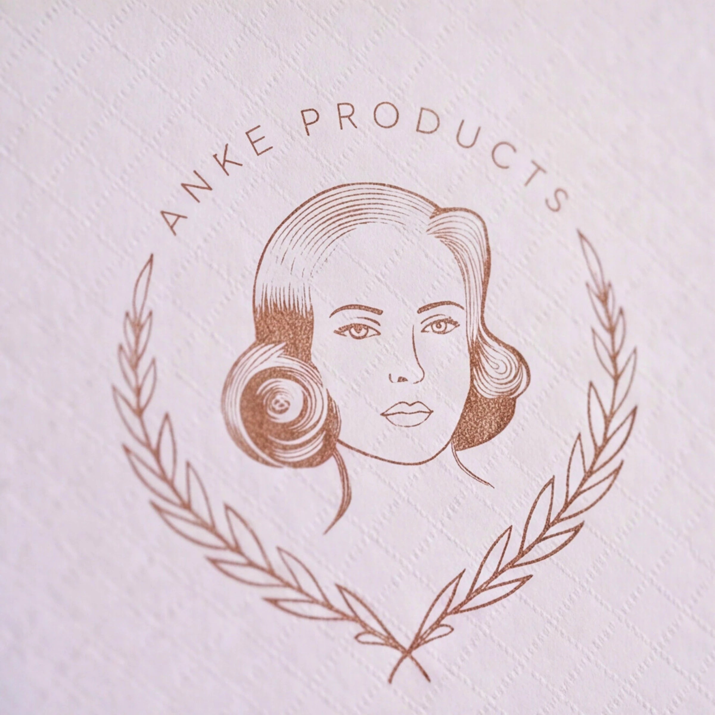 A close-up of a textured white paper surface featuring a circular embossed logo with the text 'ANKE PRODUCTS' in brown around a stylized woman's face and laurel wreath.