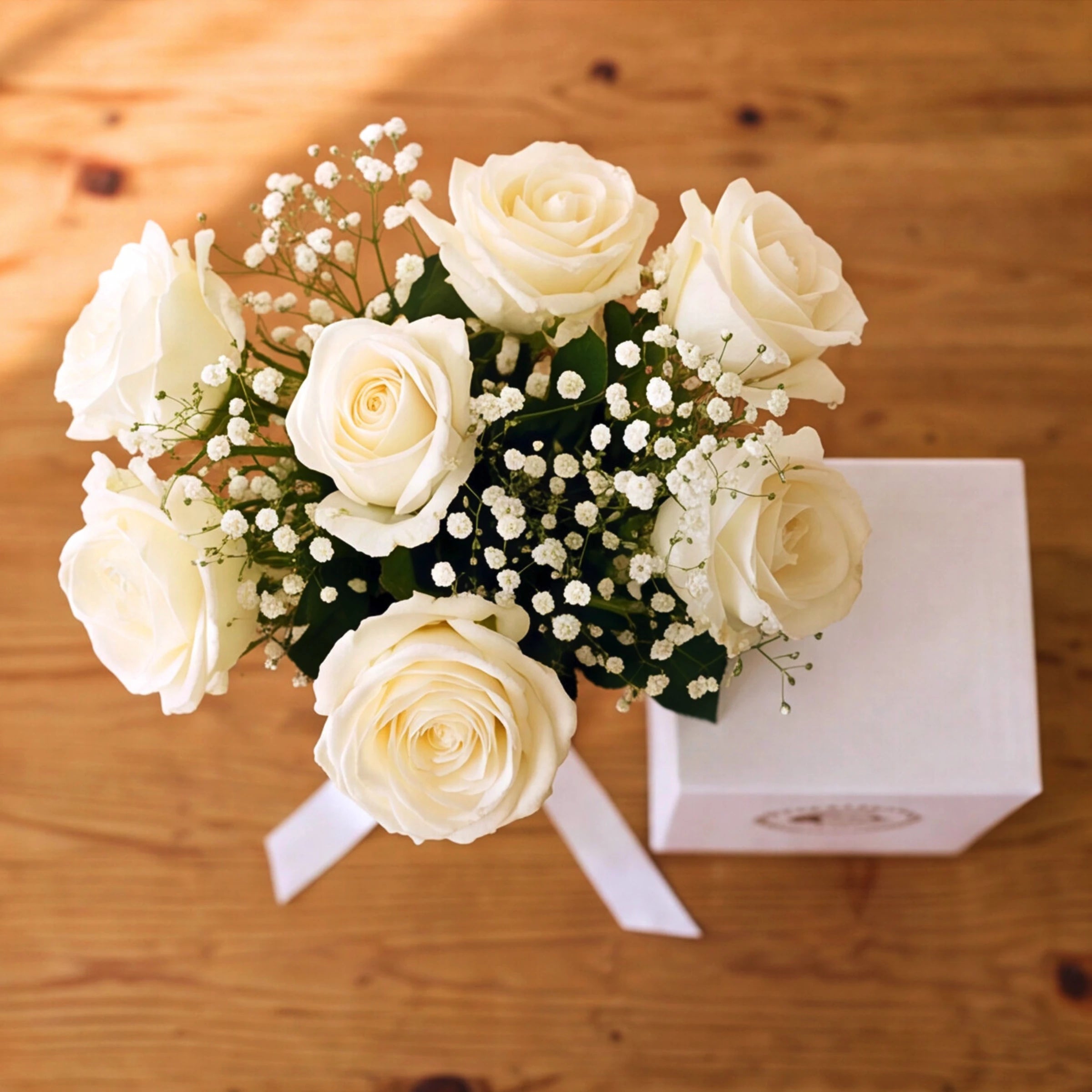 A flower arrangement of white roses and baby's breath with green leaves, tied with a white ribbon, placed on a wooden surface next to a white gift box.